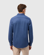 A man stands with his back to the camera against a light gray background, wearing the Psycho Bunny MENS DALTON KNIT SHIRT in elegant blue, crafted from Peruvian Pima cotton, paired with beige pants.