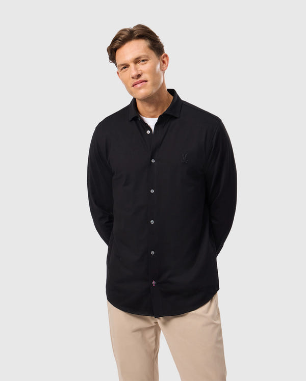 A man stands against a plain background, wearing the MENS ARTHUR JERSEY SHIRT by Psycho Bunny, a comfortable black button-up made of Pima cotton, layered over a white undershirt and paired with beige pants. He has short brown hair and is looking at the camera with a slight smile, his arms relaxed by his sides.