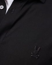 A close-up of the Psycho Bunny MENS ARTHUR JERSEY SHIRT - B6C748D200 reveals its soft Pima cotton fabric in a classic black color, complete with an embroidered bunny and crossbones logo on the chest. This comfortable shirt is designed with a buttoned collar and white buttons at the top, blending style and luxury effortlessly.