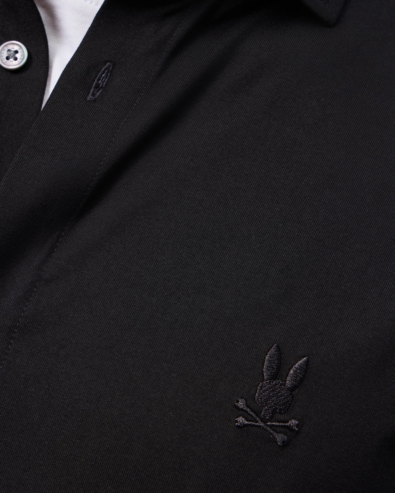 A close-up of the Psycho Bunny MENS ARTHUR JERSEY SHIRT - B6C748D200 reveals its soft Pima cotton fabric in a classic black color, complete with an embroidered bunny and crossbones logo on the chest. This comfortable shirt is designed with a buttoned collar and white buttons at the top, blending style and luxury effortlessly.