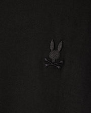 A close-up of the MENS ARTHUR JERSEY SHIRT by Psycho Bunny features a dark jersey fabric with an embroidered logo, highlighting a stylized rabbit head and crossed bones beneath, adding a distinctive design element.