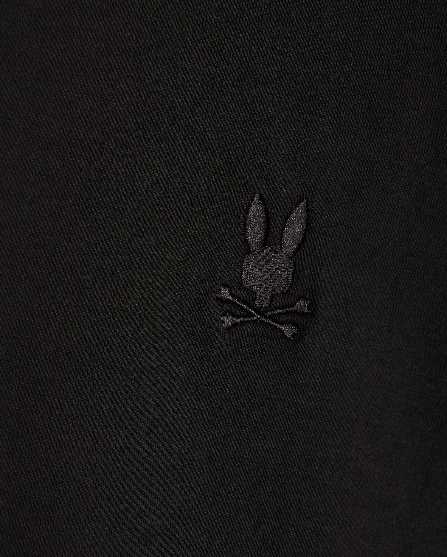 A close-up of the MENS ARTHUR JERSEY SHIRT by Psycho Bunny features a dark jersey fabric with an embroidered logo, highlighting a stylized rabbit head and crossed bones beneath, adding a distinctive design element.