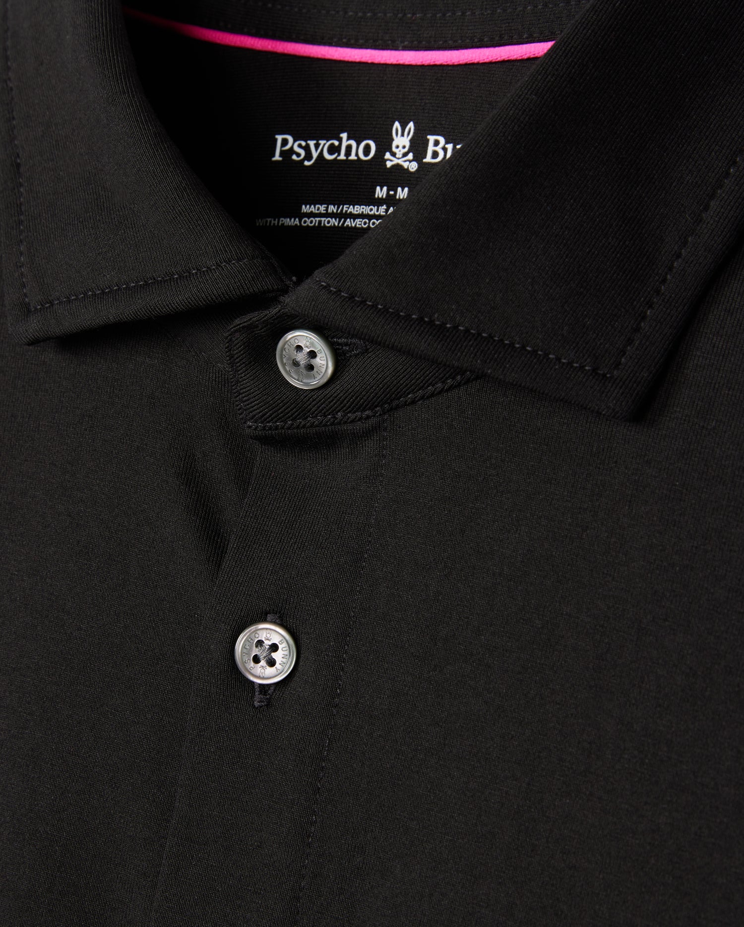 Close-up of a black MENS ARTHUR JERSEY SHIRT - B6C748D200 crafted from Pima cotton, featuring two visible buttons and a small bunny logo. The tag indicates it's from Psycho Bunny and is medium in size. The fabric is soft, smooth, and neatly stitched, epitomizing comfortable shirting.