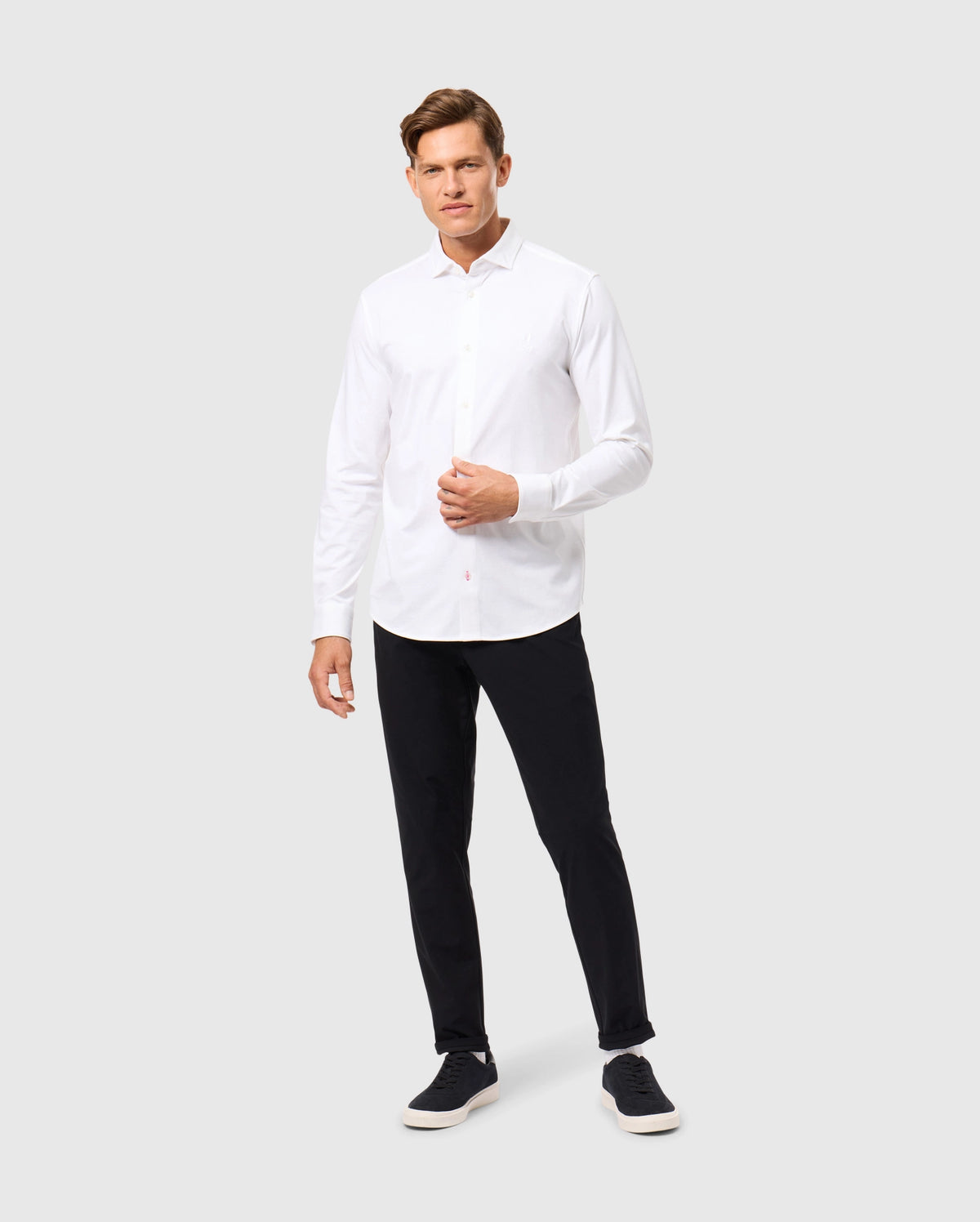 A man wearing a white Psycho Bunny MENS ARTHUR JERSEY SHIRT - B6C748D200, crafted from stretch Pima cotton, stands against a plain gray backdrop. He pairs it with black pants and black sneakers, with his right arm slightly bent and relaxed by his side.