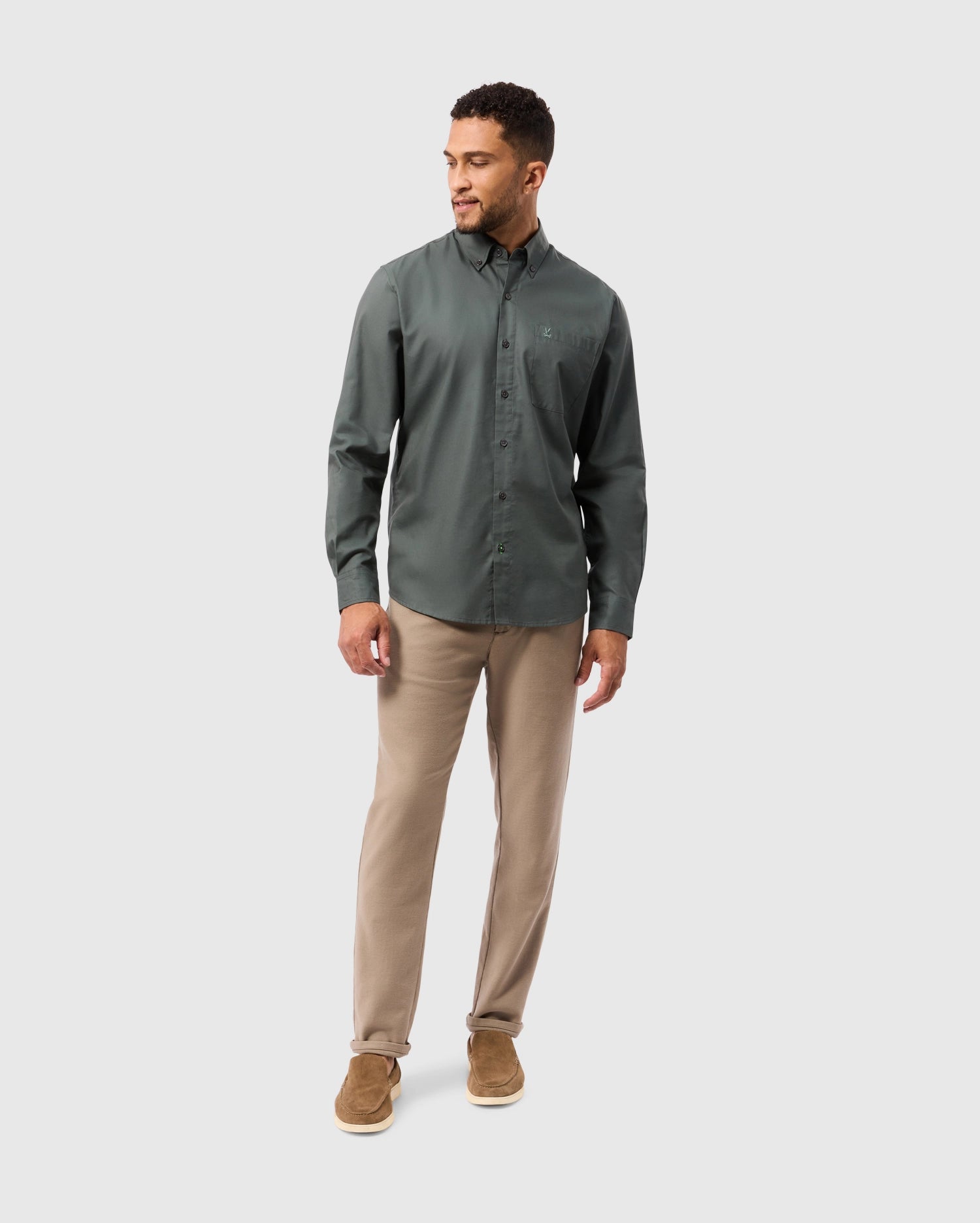 A person dressed in a dark green Psycho Bunny MENS JARED COTTON MODAL SHIRT - B6C774D200 and beige pants stands against a light gray background. They are wearing brown shoes and glancing down to their left with a slight smile.