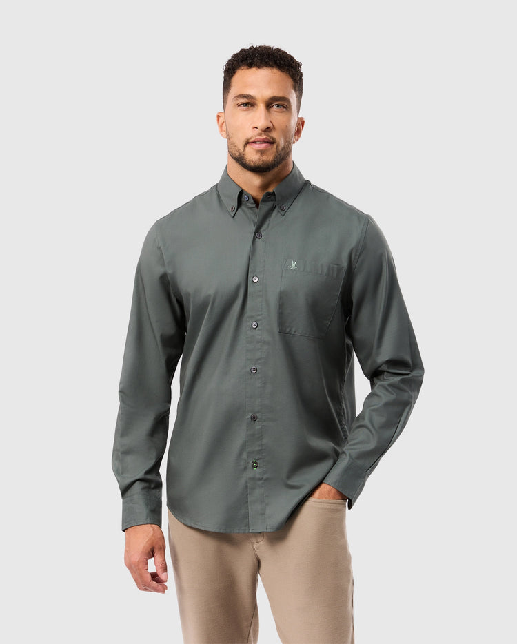 A man wearing a long-sleeve, button-up, dark green *MENS JARED COTTON MODAL SHIRT - B6C774D200* by Psycho Bunny and beige pants against a plain light gray background.