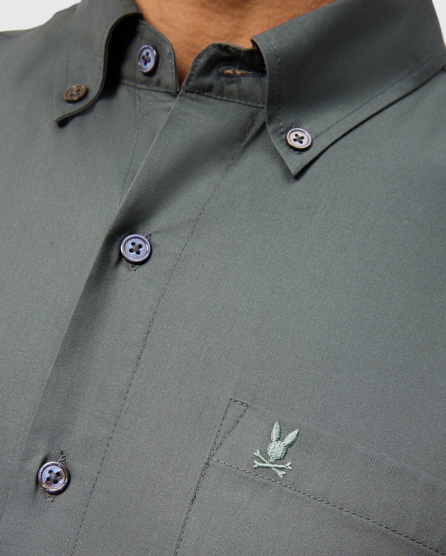 A close-up of a person wearing the Psycho Bunny MENS JARED COTTON MODAL SHIRT in dark green, featuring an embroidered logo on the chest pocket. This shirt includes a button-down collar and dark buttons.