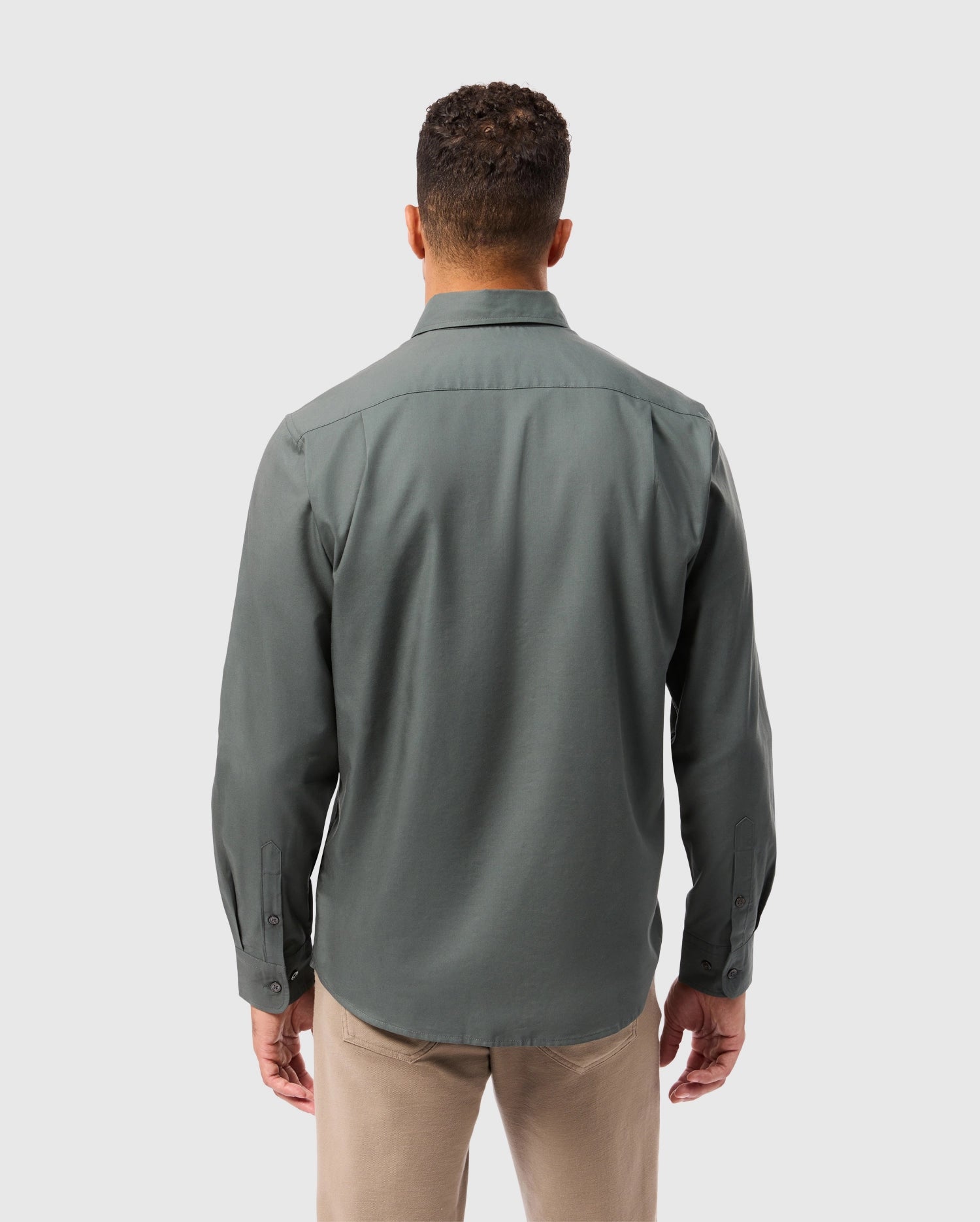 A man with short curly hair is wearing a long-sleeved, dark green Mens Jared Cotton Modal Shirt - B6C774D200 by Psycho Bunny and beige pants. He is facing away from the camera, standing against a light gray background.