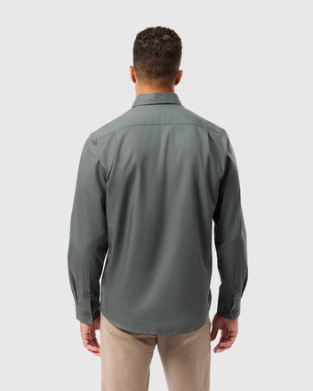 A man with short curly hair is wearing a long-sleeved, dark green Mens Jared Cotton Modal Shirt - B6C774D200 by Psycho Bunny and beige pants. He is facing away from the camera, standing against a light gray background.