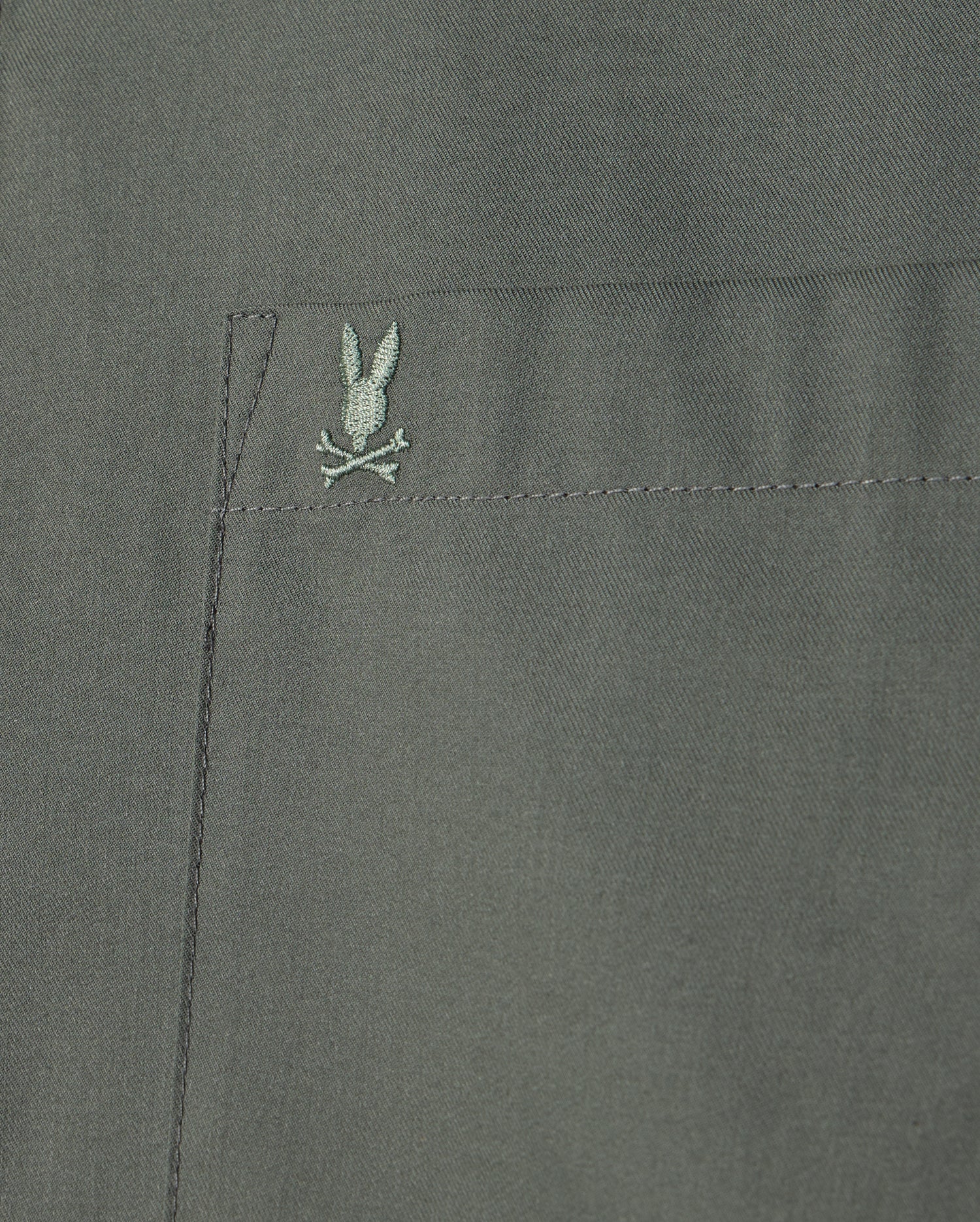 Close-up of the Psycho Bunny MENS JARED COTTON MODAL SHIRT, highlighting a pocket with an embroidered rabbit and crossbones motif in green on a muted green background.