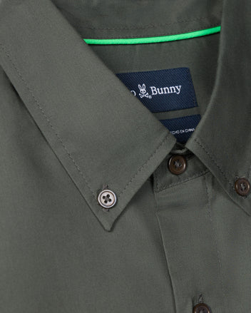 Close-up of the MENS JARED COTTON MODAL SHIRT in dark green, featuring a button-down collar. A clothing tag with Psycho Bunny's signature subtle bunny logo is visible, complemented by a thin green strip inside the collar. Crafted from premium cotton, this piece offers both style and comfort.