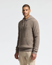 A man stands against a plain white background wearing Psycho Bunny's MENS LEVAN WAFFLE HOODED SWEATER (B6E246E200) in beige cashmere-wool, featuring an embroidered chest bunny. He's dressed in light pants, with short hair and a neutral expression, his hands by his sides.