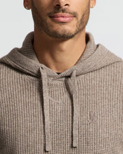 A man with a short beard and small hoop earrings is wearing the Psycho Bunny MENS LEVAN WAFFLE HOODED SWEATER (B6E246E200), a textured beige cashmere-wool hoodie with drawstrings featuring an embroidered bunny logo on the chest. The background is plain and light-colored.