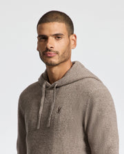 A person with short hair wearing a Psycho Bunny MENS LEVAN WAFFLE HOODED SWEATER - B6E246E200 in beige stands against a plain white background, looking slightly towards the camera.