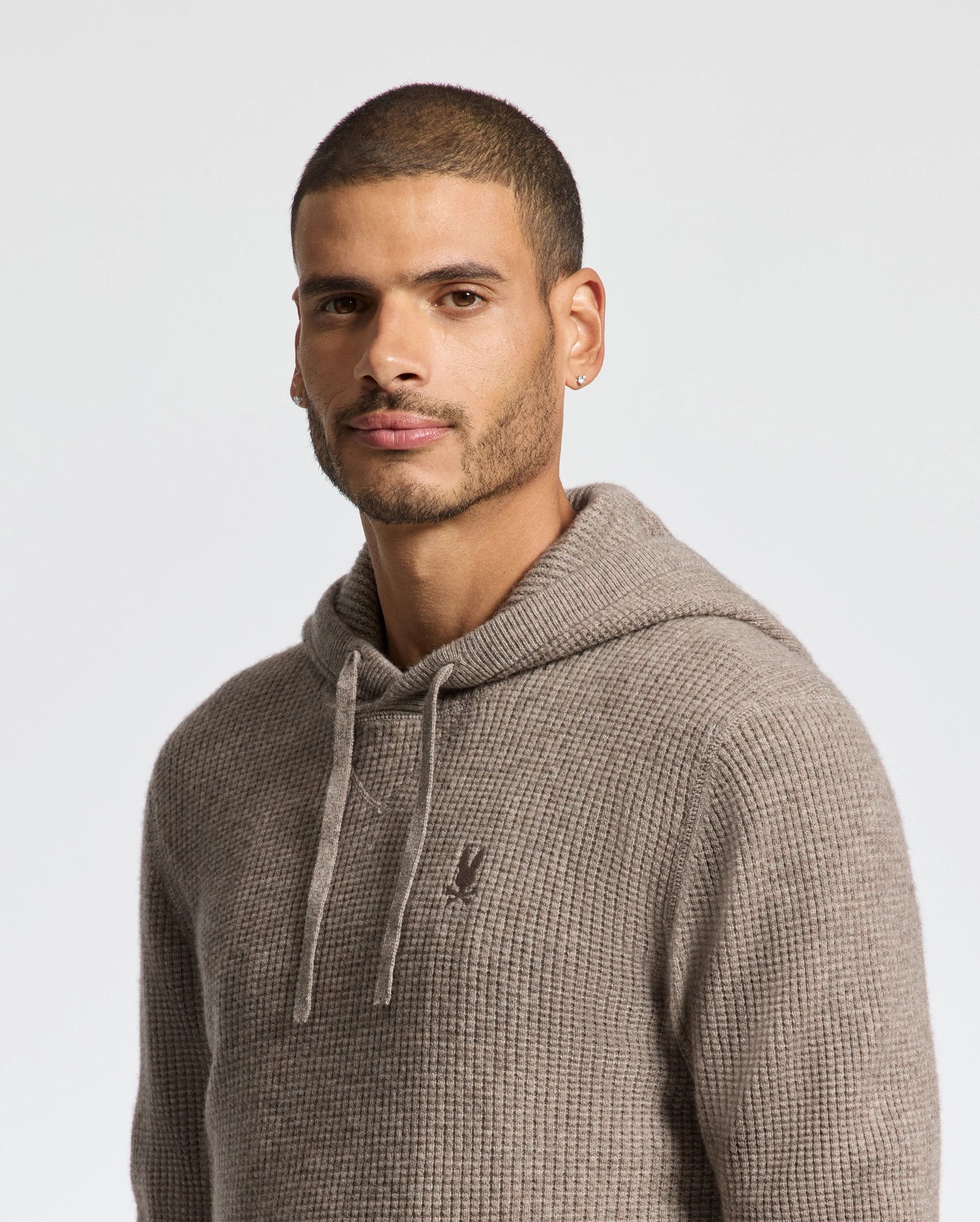 A person with short hair wearing a Psycho Bunny MENS LEVAN WAFFLE HOODED SWEATER - B6E246E200 in beige stands against a plain white background, looking slightly towards the camera.