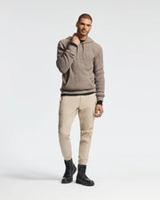 Wearing the Psycho Bunny MENS LEVAN WAFFLE HOODED SWEATER in brown, paired with beige cargo pants and black boots, a man stands against a plain background. He touches his hoodie near the embroidered bunny at the neck, smiling subtly.
