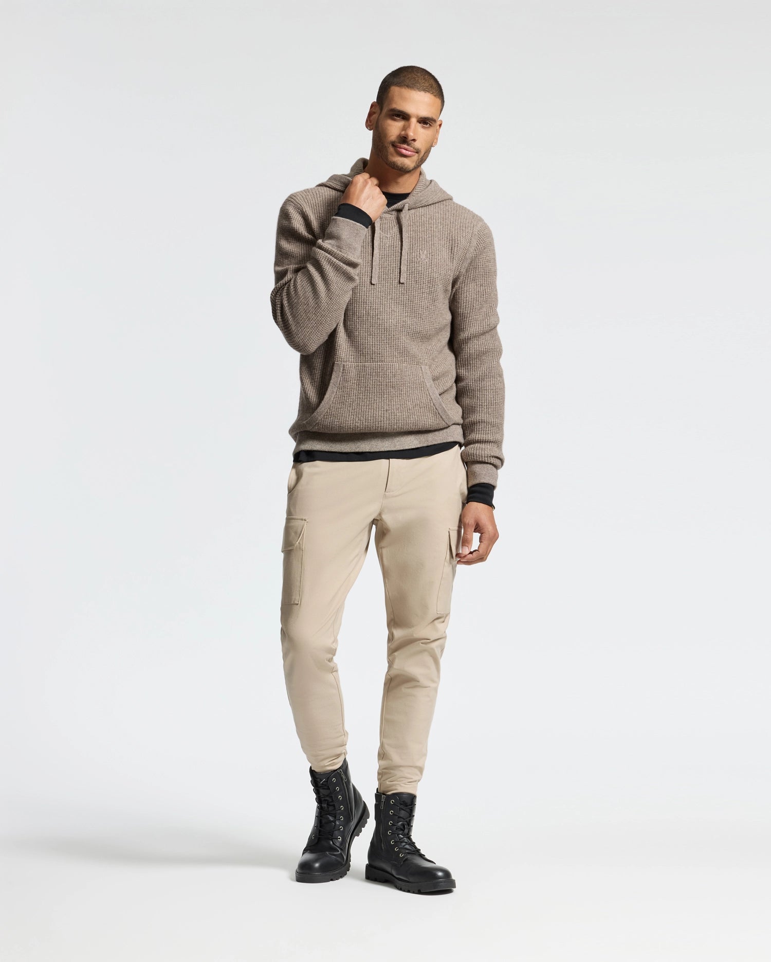 Wearing the Psycho Bunny MENS LEVAN WAFFLE HOODED SWEATER in brown, paired with beige cargo pants and black boots, a man stands against a plain background. He touches his hoodie near the embroidered bunny at the neck, smiling subtly.