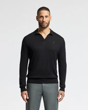 A person stands against a plain background, wearing a MENS JOHNNY COLLAR SWEATER - B6E258E200 from Psycho Bunny, crafted from a cotton-wool blend, along with gray pants. They have short hair and a beard.