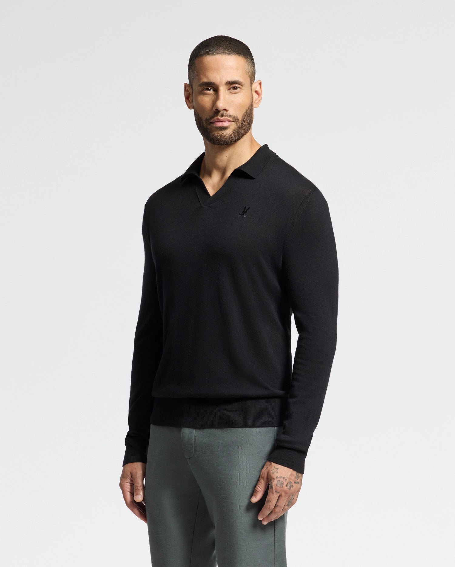 A man with a short beard poses against a plain white background, sporting the MENS JOHNNY COLLAR SWEATER - B6E258E200 by Psycho Bunny. The sleek black sweater, crafted from a comfortable cotton-wool blend, is paired with gray pants as he rests his hands by his sides.