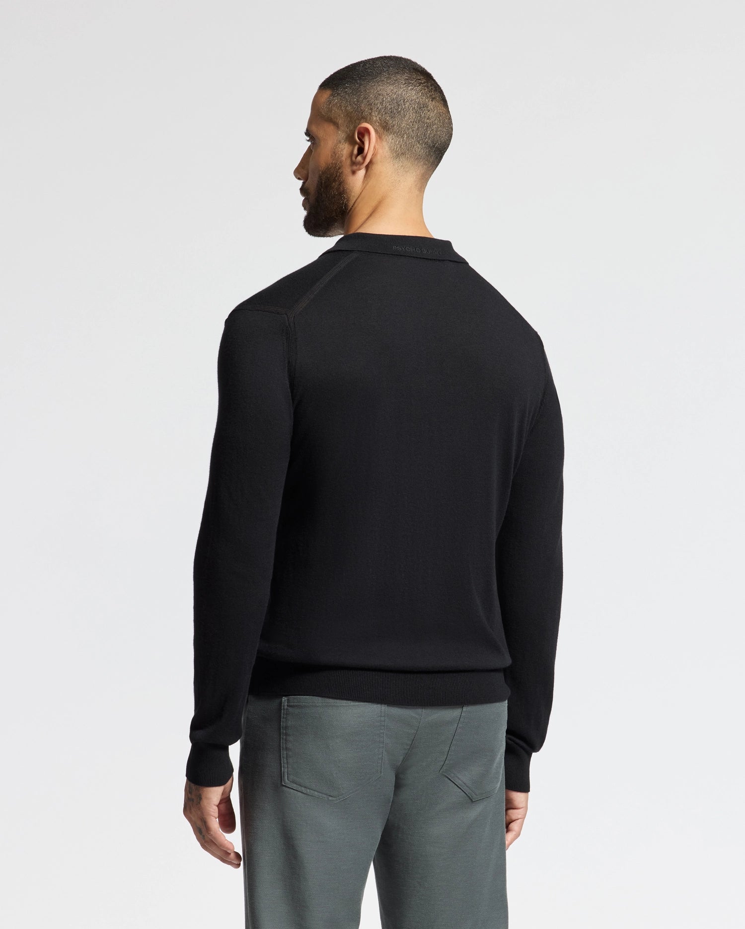A man with short hair poses against a white backdrop, dressed in the Psycho Bunny MENS JOHNNY COLLAR SWEATER - B6E258E200 in black, crafted from a comfortable cotton-wool blend, along with gray pants. His body is angled slightly to the side, emphasizing the back of his sweater and part of his profile.