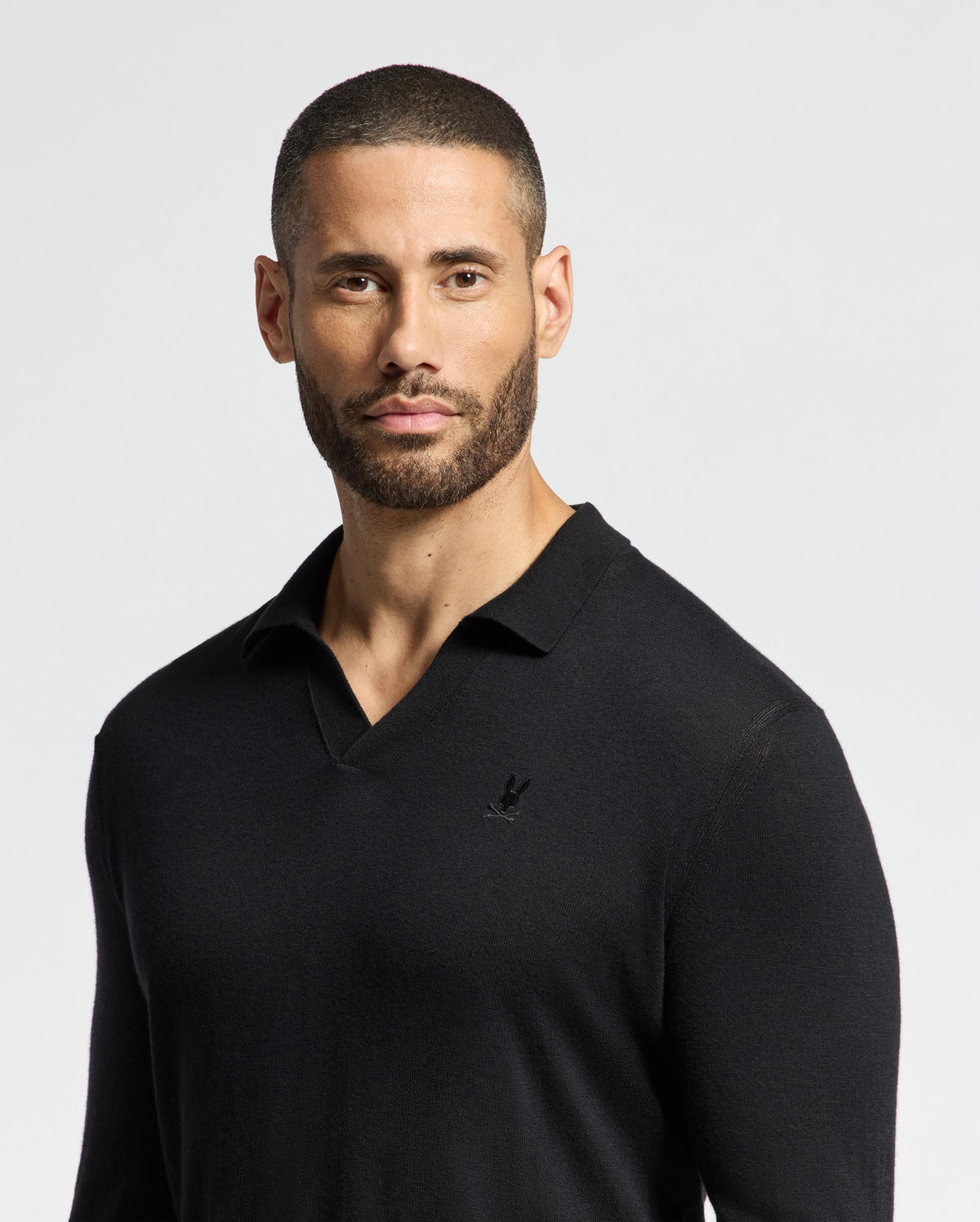 A man with a neatly trimmed beard and short hair is wearing a black Psycho Bunny Men's Johnny Collar Sweater (B6E258E200) made from a soft cotton-wool blend, featuring a small logo on the chest. He stands against a plain, light background, looking forward with a neutral expression.