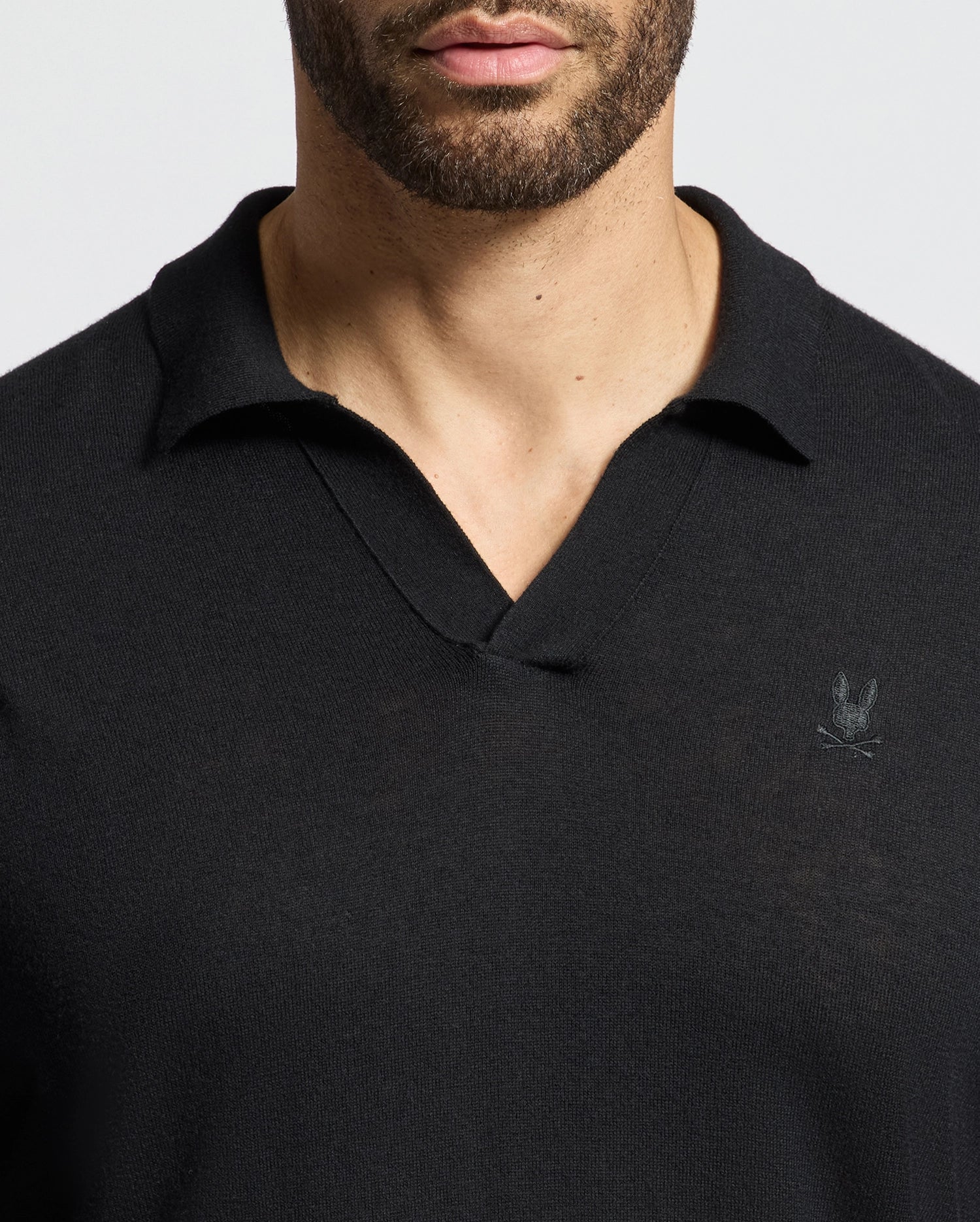A man wears the Psycho Bunny Johnny Collar sweater in black, crafted from a cotton-wool blend. It features a small embroidered emblem on the chest and a subtle v-neckline. The plain, light-colored background highlights the stylish simplicity of this MENS JOHNNY COLLAR SWEATER - B6E258E200.