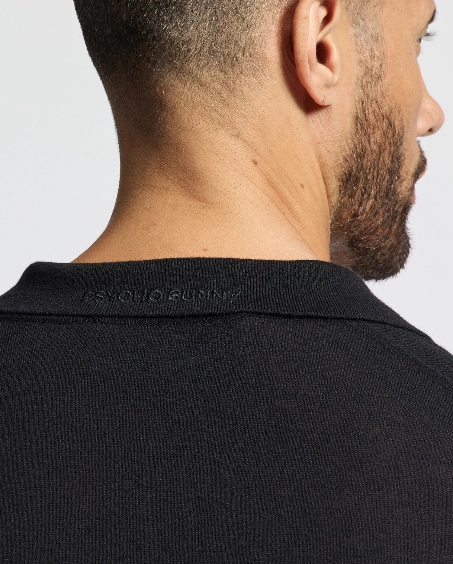 A person with short hair and a beard is shown from behind, wearing the Psycho Bunny MENS JOHNNY COLLAR SWEATER - B6E258E200 in black. The sleek look, featuring "PSYCHO BUNNY" subtly embroidered on the back of the collar, stands out against the plain white background.
