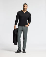A man with short hair and a beard stands against a plain background, wearing the sleek black MENS JOHNNY COLLAR SWEATER - B6E258E200 by Psycho Bunny and gray pants. He holds a black jacket in one hand, complementing his classic look with black shoes.
