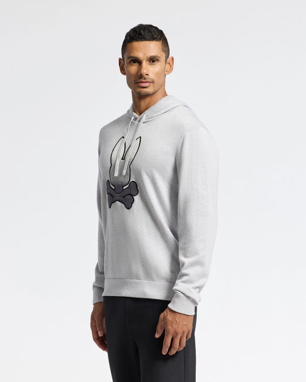 A man wearing a light gray Psycho Bunny MENS BULDER HOODED SWEATER - B6E261E200 featuring a bold black and white graphic with horns and a cross stands against a plain, light gray backdrop.