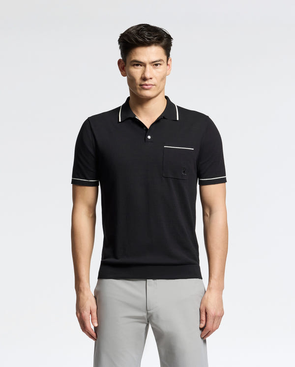 A person stands wearing the Psycho Bunny MENS LONGFORD FINE KNIT POLO - B6E500F200, a black shirt with white trim and an embroidered chest pocket, paired with light gray pants. The fine-knit cotton polo complements the plain white background seamlessly.