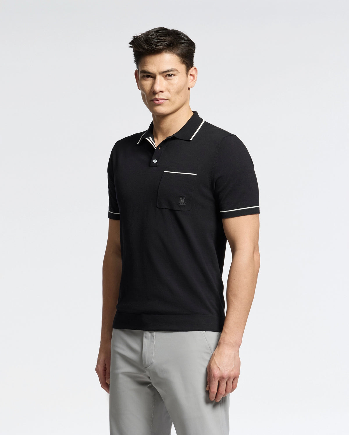 A man in a Psycho Bunny MENS LONGFORD FINE KNIT POLO - B6E500F200 with white trim and light gray pants stands against a white background, exuding elegance as he gazes neutrally at the camera.