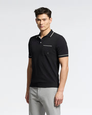 A man in a Psycho Bunny MENS LONGFORD FINE KNIT POLO - B6E500F200 with white trim and light gray pants stands against a white background, exuding elegance as he gazes neutrally at the camera.