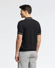 A person with short dark hair, facing away, dons a Psycho Bunny MENS LONGFORD FINE KNIT POLO in black with white trim and light gray pants. The fine-knit cotton enhances the minimalist style against a plain white backdrop.