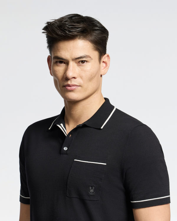 A person with short dark hair wears a black Psycho Bunny Longford Fine Knit Polo, featuring a white-trimmed collar and an embroidered chest pocket, set against a plain gray background.