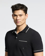A person with short dark hair wears a black Psycho Bunny Longford Fine Knit Polo, featuring a white-trimmed collar and an embroidered chest pocket, set against a plain gray background.