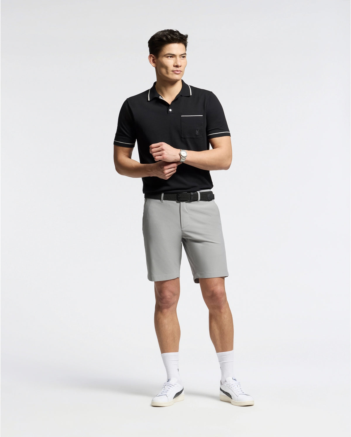 A man wears a black Psycho Bunny MENS LONGFORD FINE KNIT POLO shirt with an embroidered chest pocket, paired with gray shorts, white socks, and white sneakers. He stands casually against a plain white background adjusting the watch on his wrist.