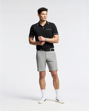A man wears a black Psycho Bunny MENS LONGFORD FINE KNIT POLO shirt with an embroidered chest pocket, paired with gray shorts, white socks, and white sneakers. He stands casually against a plain white background adjusting the watch on his wrist.