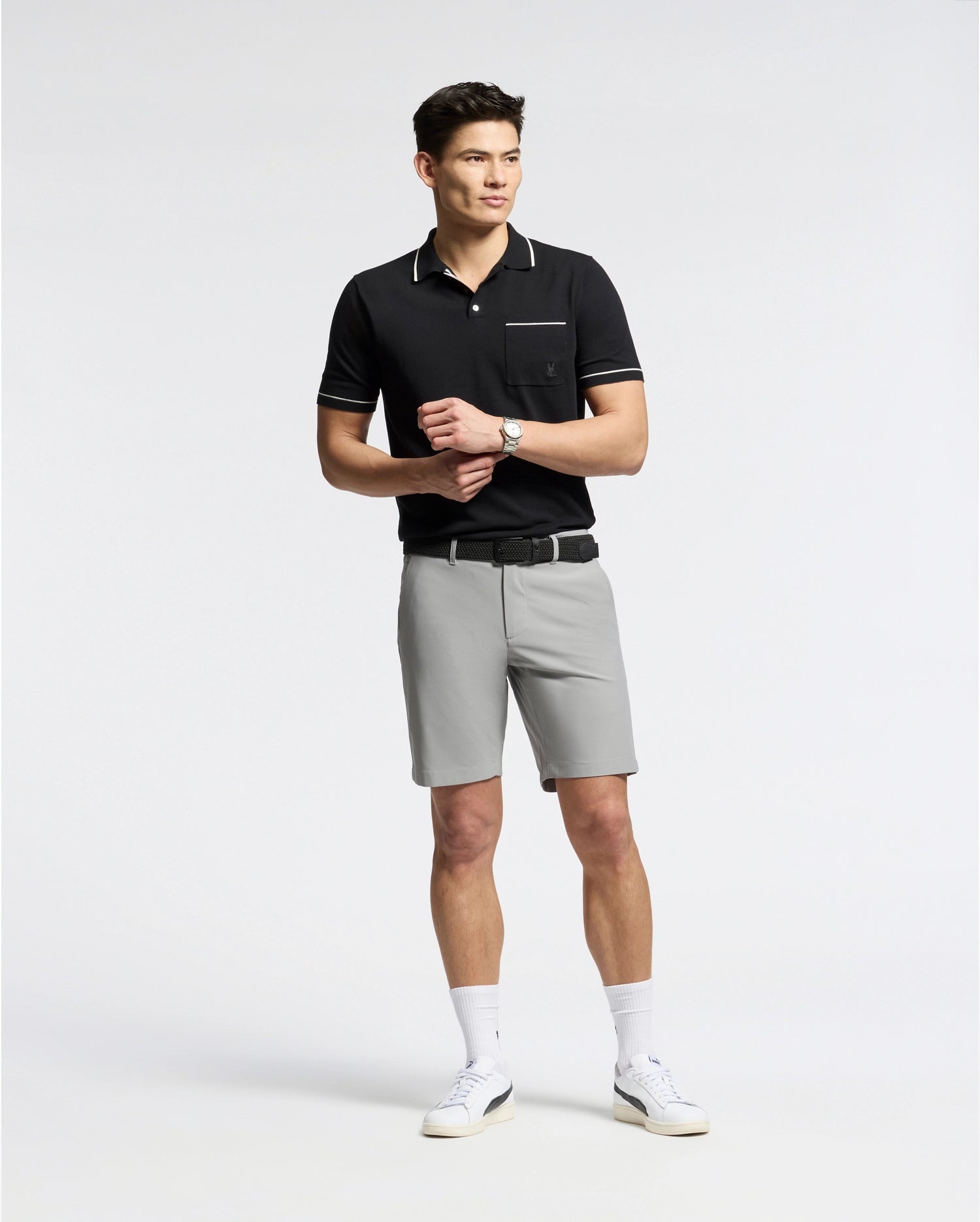 A man wears a black Psycho Bunny MENS LONGFORD FINE KNIT POLO shirt with an embroidered chest pocket, paired with gray shorts, white socks, and white sneakers. He stands casually against a plain white background adjusting the watch on his wrist.