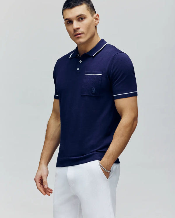 A man wears a Psycho Bunny MENS LONGFORD FINE KNIT POLO (B6E500F200) in slim fit navy blue with white trim and white pants, standing against a light gray background and looking at the camera with a neutral expression.