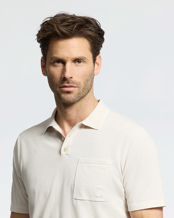 A man with short brown hair and light stubble wears a cream-colored Psycho Bunny MENS LONGFORD FINE KNIT POLO - B6E500F200, featuring an embroidered Bunny on the chest pocket. He stands against a plain light background.