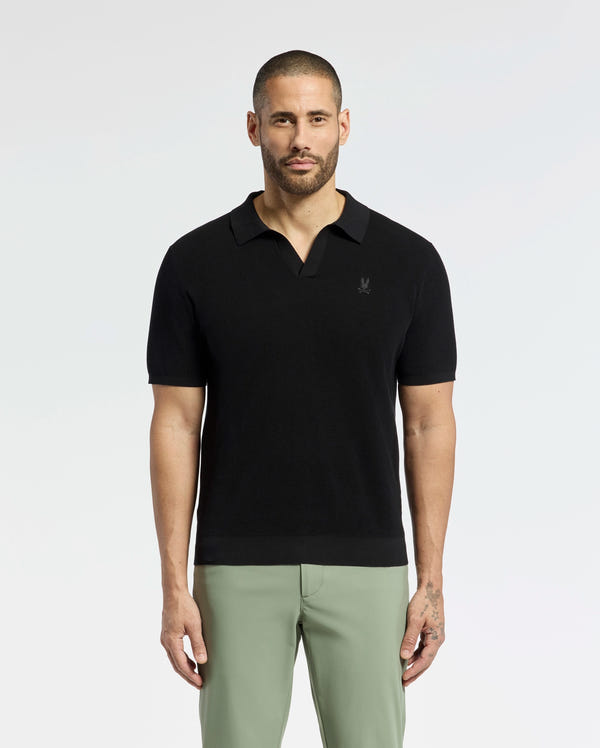 A bearded man confidently wears a breathable Psycho Bunny MENS LEETON JOHNNY COLLAR KNIT POLO (B6E562F200) in black, paired with light green pants. With an embroidered bunny, the 100% cotton shirt fits him perfectly as he gazes directly at the camera, showcasing his left-hand tattoo against a plain backdrop.