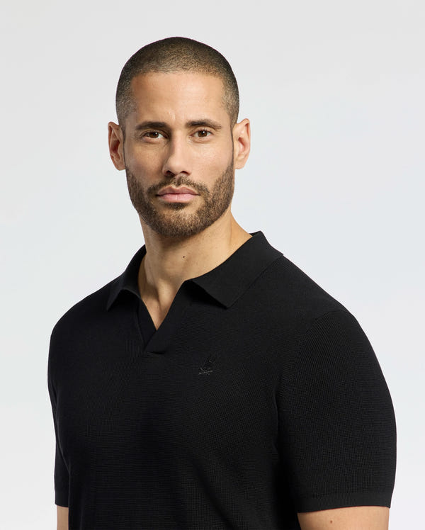 A bearded, short-haired person confidently looks at the camera while wearing a Psycho Bunny MENS LEETON JOHNNY COLLAR KNIT POLO, made of breathable black 100% cotton with an embroidered Bunny. The background is plain and light-colored.