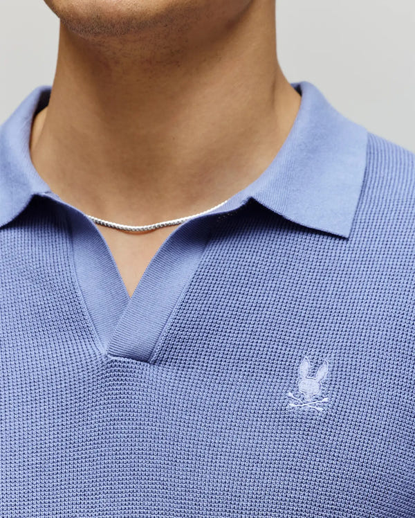 A person wears the Psycho Bunny MENS LEETON KNIT POLO SHIRT (B6E562F200), a textured light blue sweater polo with a Johnny collar and small white bunny logo, paired with a thin silver chain; only their lower face, neck, and upper chest are visible.