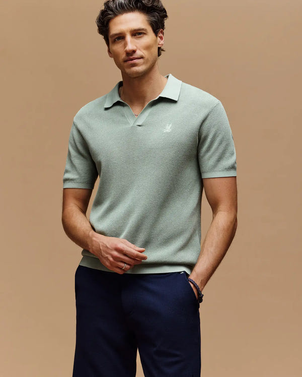 A man with wavy brown hair wears a Psycho Bunny MENS LEETON JOHNNY COLLAR KNIT POLO (B6E562F200) in light green and dark navy pants, standing against a beige background, facing the camera with a relaxed expression.