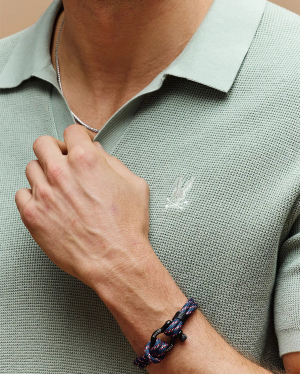 Wearing the Psycho Bunny MENS LEETON JOHNNY COLLAR KNIT POLO (B6E562F200) in light green with an embroidered Bunny logo, a silver chain necklace, and a blue and black bracelet for a chic, casual style.