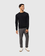 A person wearing the Psycho Bunny MENS BELMORE EXTRA FINE COTTON SWEATER in black, featuring a tonal embroidered Bunny logo, is paired with gray pants and white sneakers against a plain light gray background. Made from extrafine cotton, the outfit showcases premium stitching as they smile and look to the side.