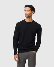A person wearing the Psycho Bunny MENS BELMORE EXTRA FINE COTTON SWEATER - B6E832D200, featuring an embroidered bunny logo, paired with gray pants, stands against a plain light gray background. The ensemble highlights premium stitching and is crafted from extrafine cotton. They have a neutral expression with their hands in their pockets.