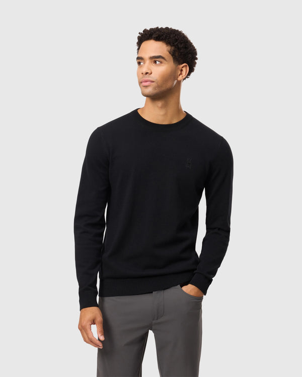 A person wearing the Psycho Bunny MENS BELMORE EXTRA FINE COTTON SWEATER - B6E832D200, featuring an embroidered bunny logo, paired with gray pants, stands against a plain light gray background. The ensemble highlights premium stitching and is crafted from extrafine cotton. They have a neutral expression with their hands in their pockets.