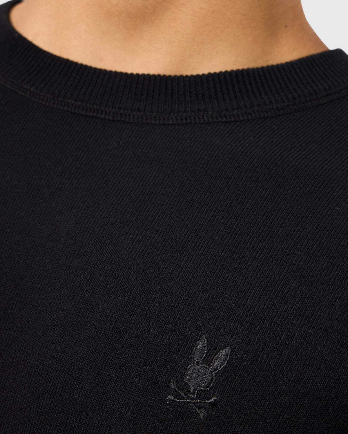 The image showcases a person wearing the Psycho Bunny MENS BELMORE EXTRA FINE COTTON SWEATER - B6E832D200, which features an embroidered bunny skull and crossbones logo on the chest. Made from extrafine cotton, this black sweater includes a ribbed neckline that highlights its premium stitching and stylish design details.