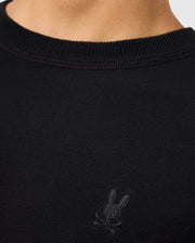 The image showcases a person wearing the Psycho Bunny MENS BELMORE EXTRA FINE COTTON SWEATER - B6E832D200, which features an embroidered bunny skull and crossbones logo on the chest. Made from extrafine cotton, this black sweater includes a ribbed neckline that highlights its premium stitching and stylish design details.